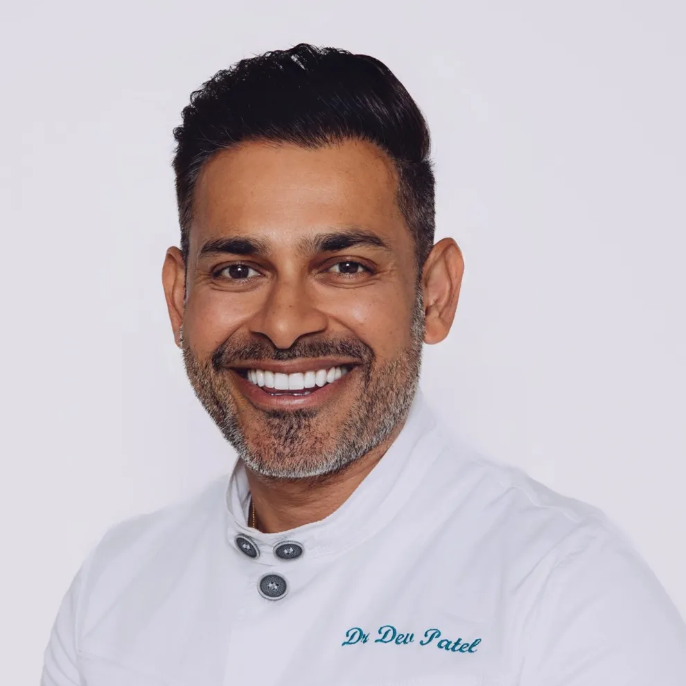 Professional headshot of Dr Dev Patel smiling in a white clinical uniform against a clean light background.